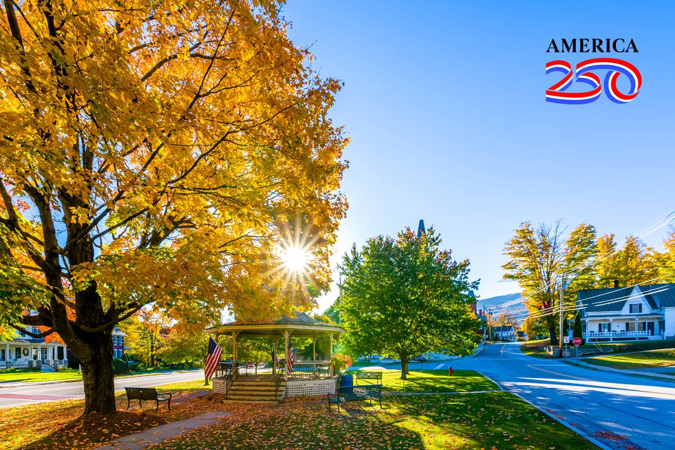 A scenic autumn street featuring vibrant yellow trees, a gazebo, benches, and American flags, with clear blue skies above.