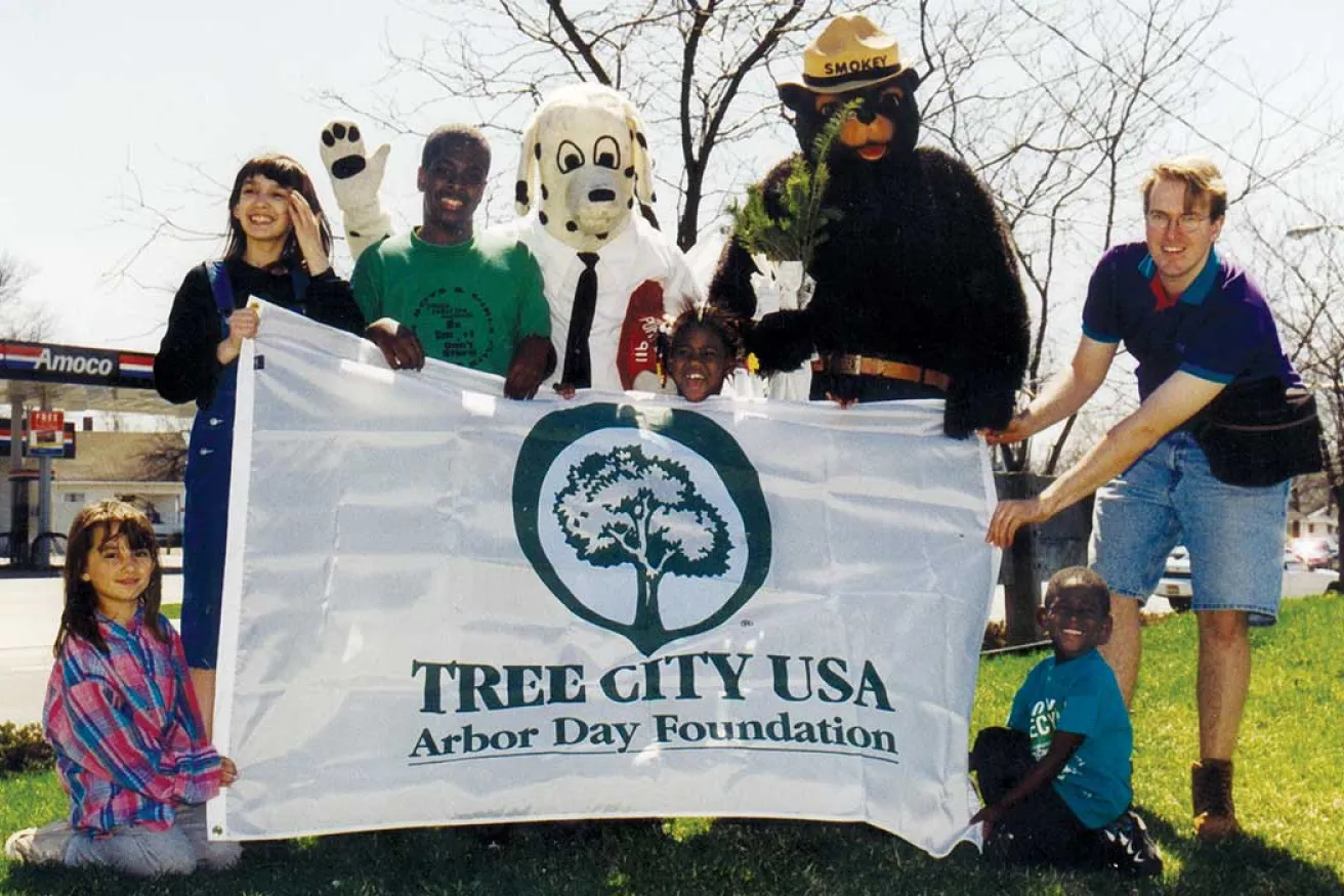 A group holds a banner for Arbor Day, featuring mascot characters, while promoting tree conservation in a park setting.