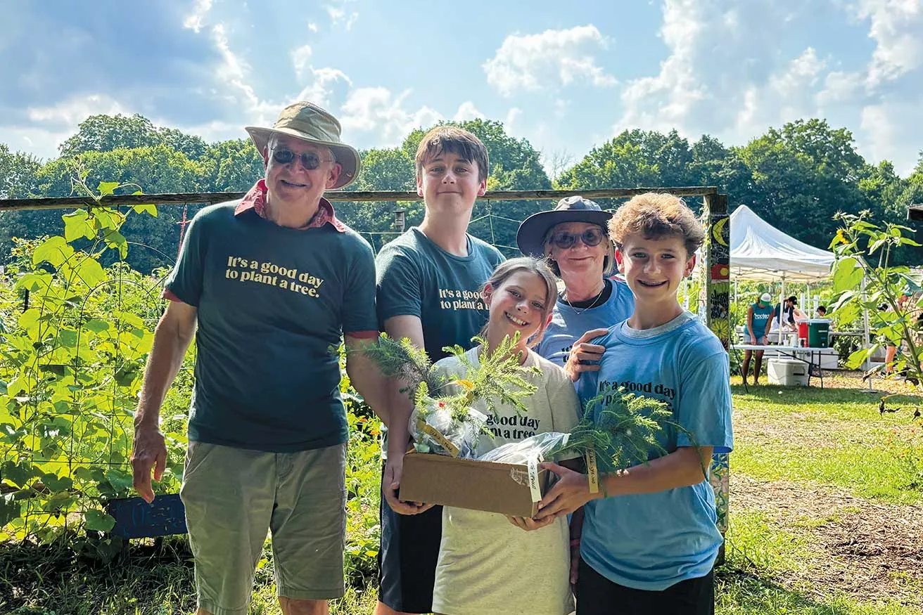 Five family members pose at an outdoor tree-planting event, wearing “It’s a good day to plant a tree” shirts and holding a box of young evergreen seedlings under a sunny sky.