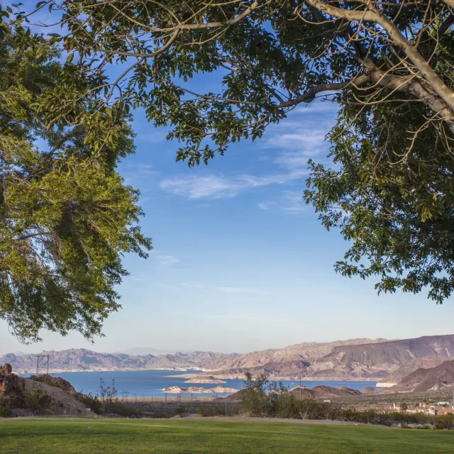 View of a serene lake surrounded by mountains, framed by lush trees and a clear blue sky, creating a tranquil landscape scene.