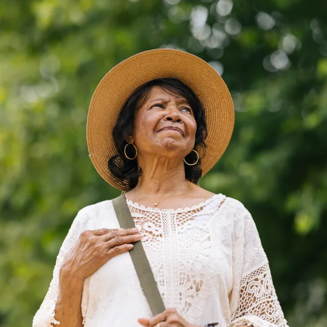 An elegantly dressed woman wearing a wide-brimmed straw hat and a white lace top stands against a lush green background.