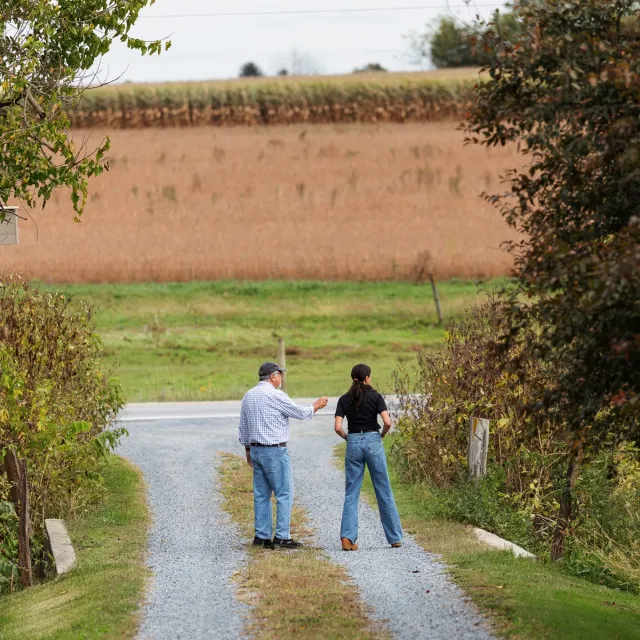 Two people stand on a gravel road surrounded by lush greenery and a field of tall crops under a cloudy sky.