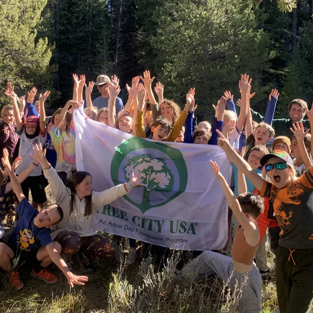 A group of children joyfully raises their arms in celebration while holding a "Tree City USA" flag in a forested area.