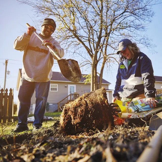 Two people work together outdoors, one using a shovel while the other holds a bag of soil near a tree stump in a sunny yard.