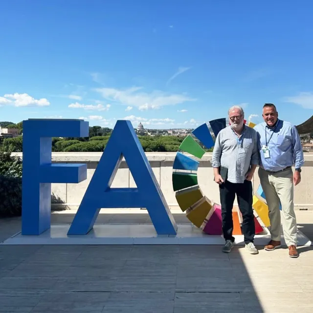 Two men stand beside large blue letters spelling "FAO," with a colorful circular emblem and a scenic view of a city under a clear sky.