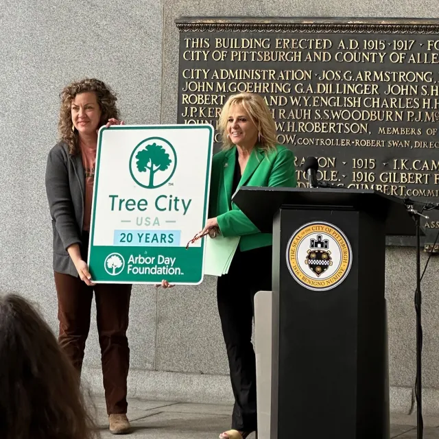 Two women hold a "Tree City USA" sign celebrating 20 years, with a backdrop of a historical plaque in Pittsburgh.
