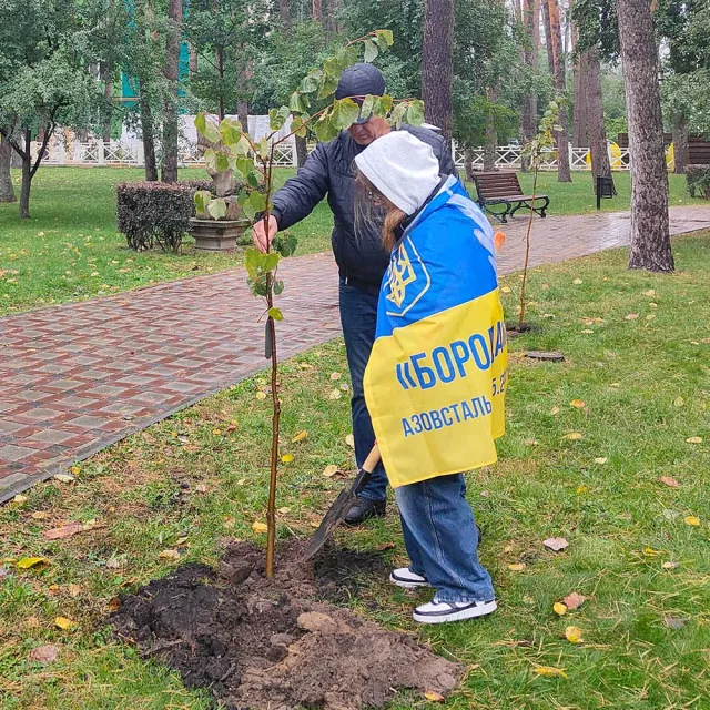 Two individuals are planting a small tree in a green park, one wearing a flag as a cape, surrounded by nature.