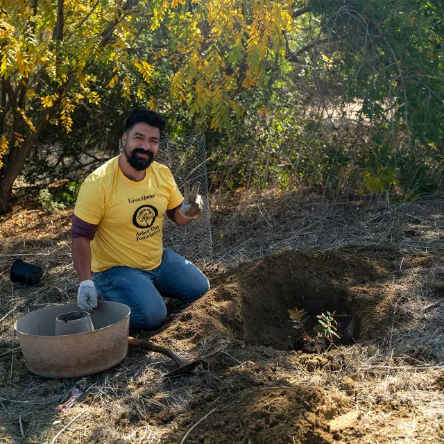 Person wearing an Arbor Day Foundation shirt kneels by a planted sapling, holding up a peace sign in a sunny wooded area.