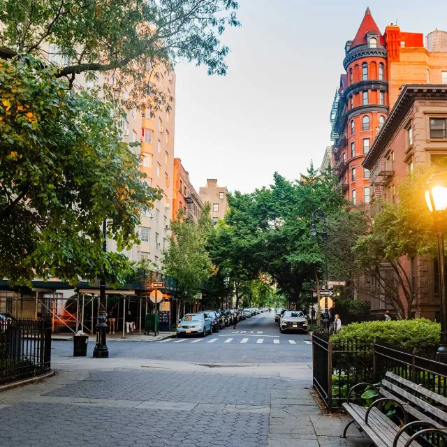 A city street featuring a bench beside a lamp post, surrounded by buildings and greenery.