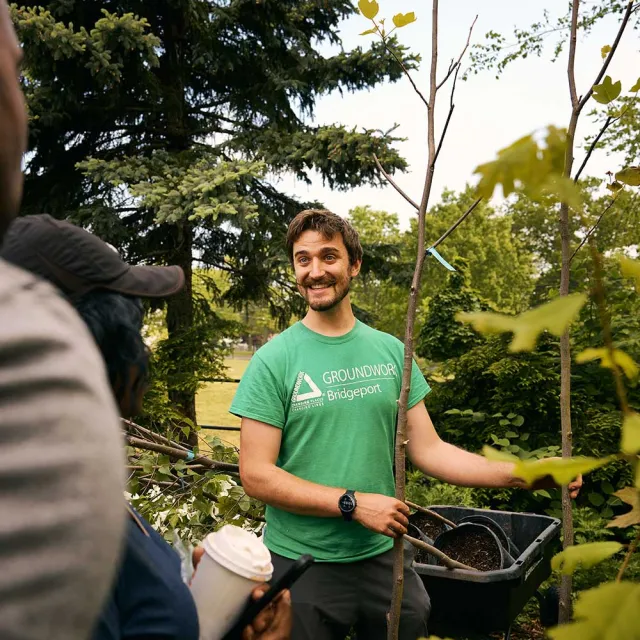 A man talking to people while holding a tree surrounded by trees outside