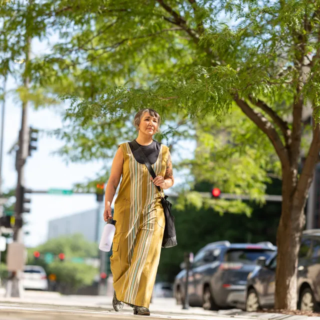 Person in yellow striped dress walking on sunny street holding a water bottle, with trees and cars in the background.