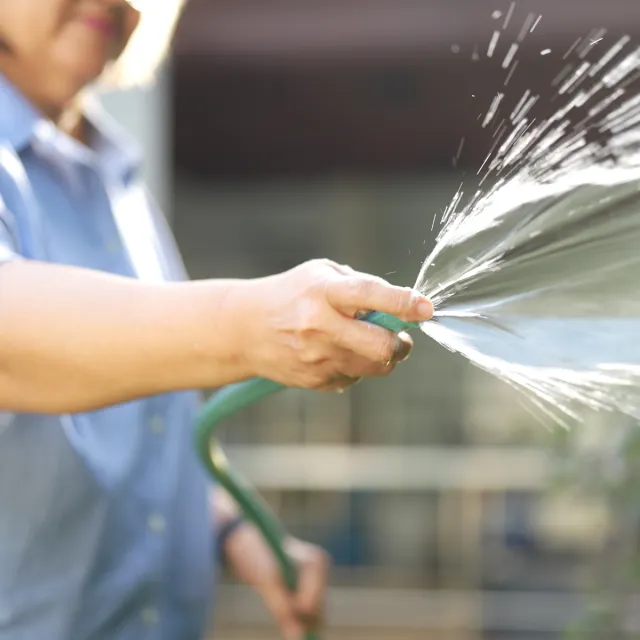 Close-up of a person watering outdoors with a green garden hose, sunlight catching the spray of water as it splashes forward.