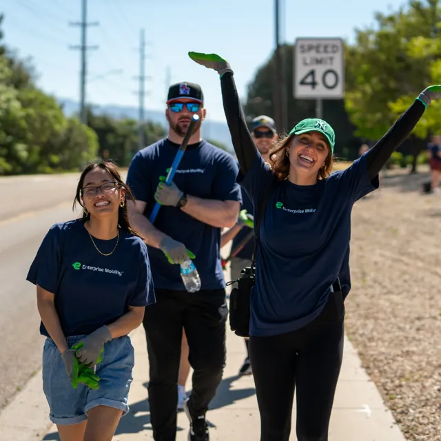A group of volunteers in matching t-shirts cleans a roadside, with one person joyfully raising their arms near a speed limit sign.