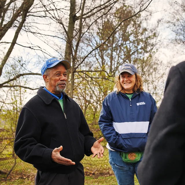Two people wearing jackets and caps chat outdoors in a wooded area during a cool day.