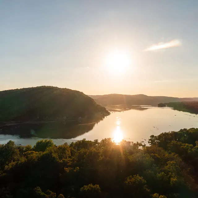 Golden sunset over a calm lake winding between wooded hills, with sunlight reflecting on the water and casting a serene, warm glow.