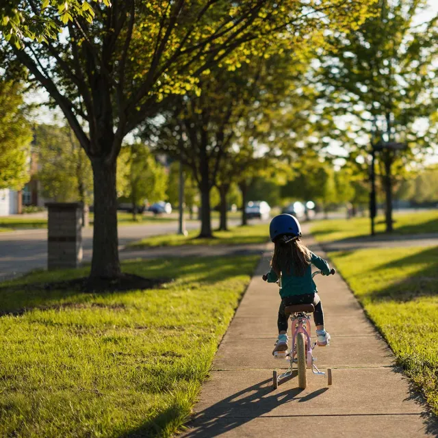 A young girl joyfully rides her bike along a sidewalk, surrounded by greenery and sunlight.