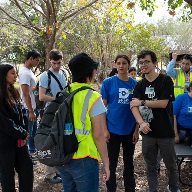 A person in a yellow jacket talking with a group of people standing outside surrounded by trees