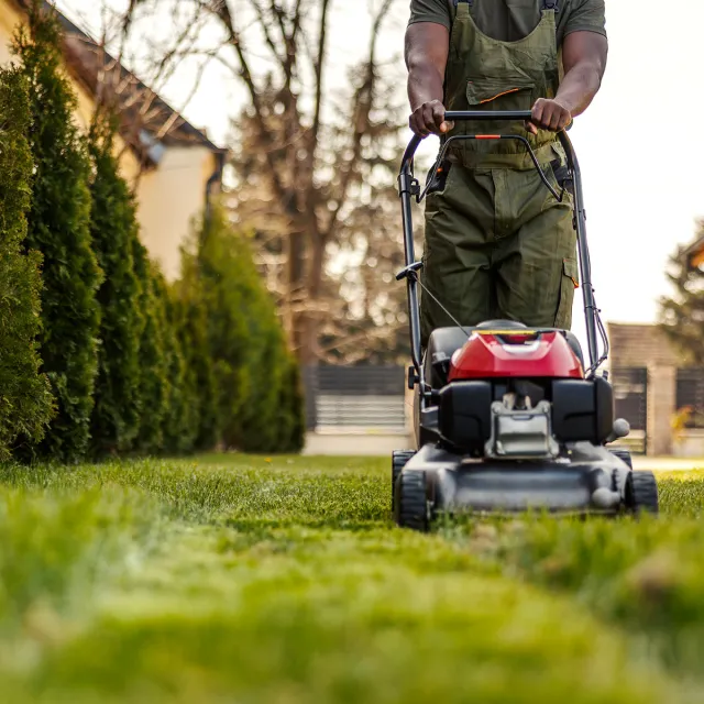A man using a lawn mower to cut grass in a residential yard on a sunny day.