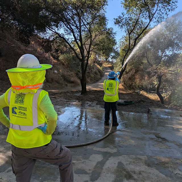Two men in safety vests spray water on a road, likely for maintenance or dust control purposes.