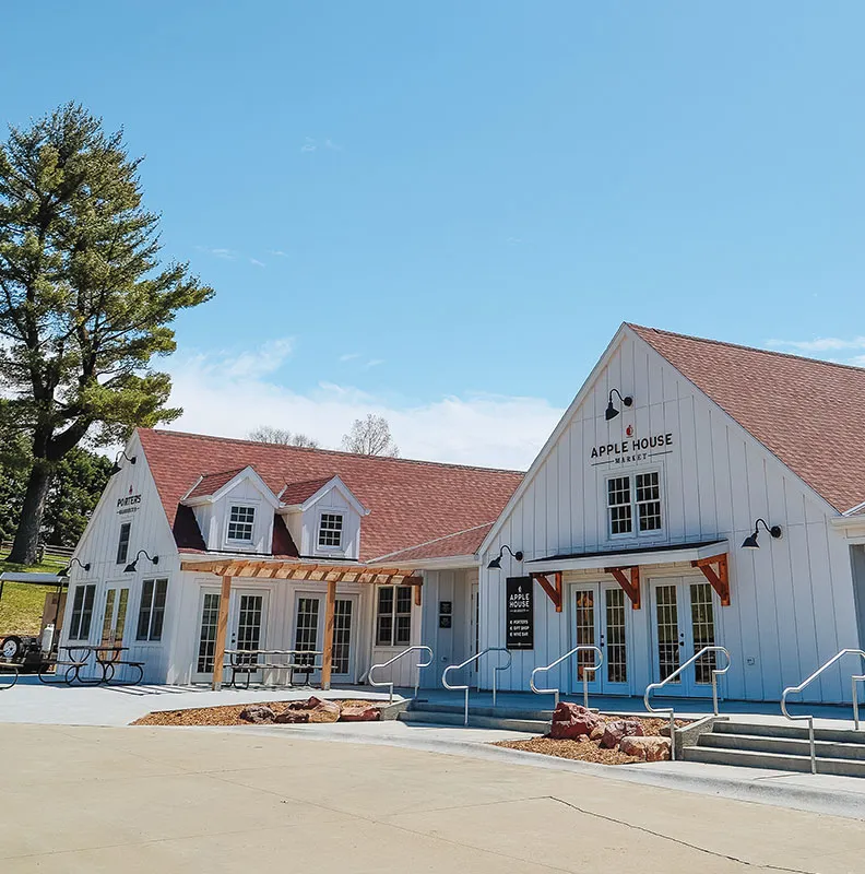 Exterior view of the Apple House Market, a charming white building with a red roof, surrounded by trees and a clear blue sky.