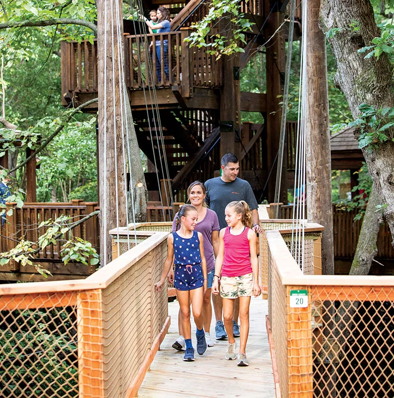 A family walks along a wooden bridge in a forest setting, surrounded by trees and rustic structures in the background.