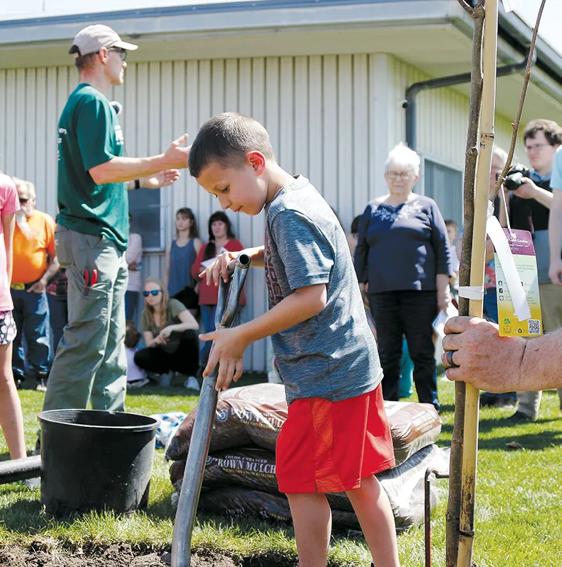 A young boy in a gray shirt and red shorts holds a gardening tool amid a crowd at a planting event near a building.