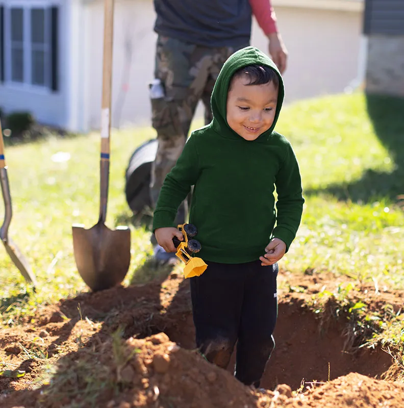 A child in a green hoodie holds a toy truck while standing near a freshly dug hole in the yard, with gardening tools in the background.