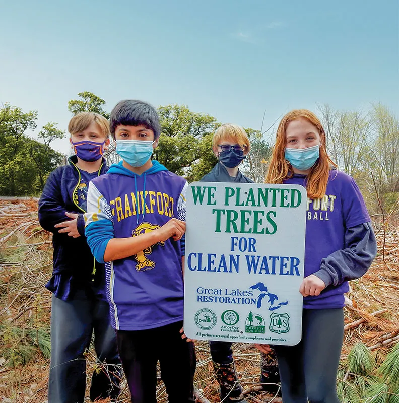 A group of four children stands proudly beside a sign reading "We planted trees for clean water," surrounded by a wooded area.