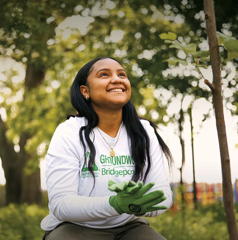 A woman in a white long-sleeve shirt and green gloves sits outdoors among trees, appearing thoughtful at a tree in a serene park setting.