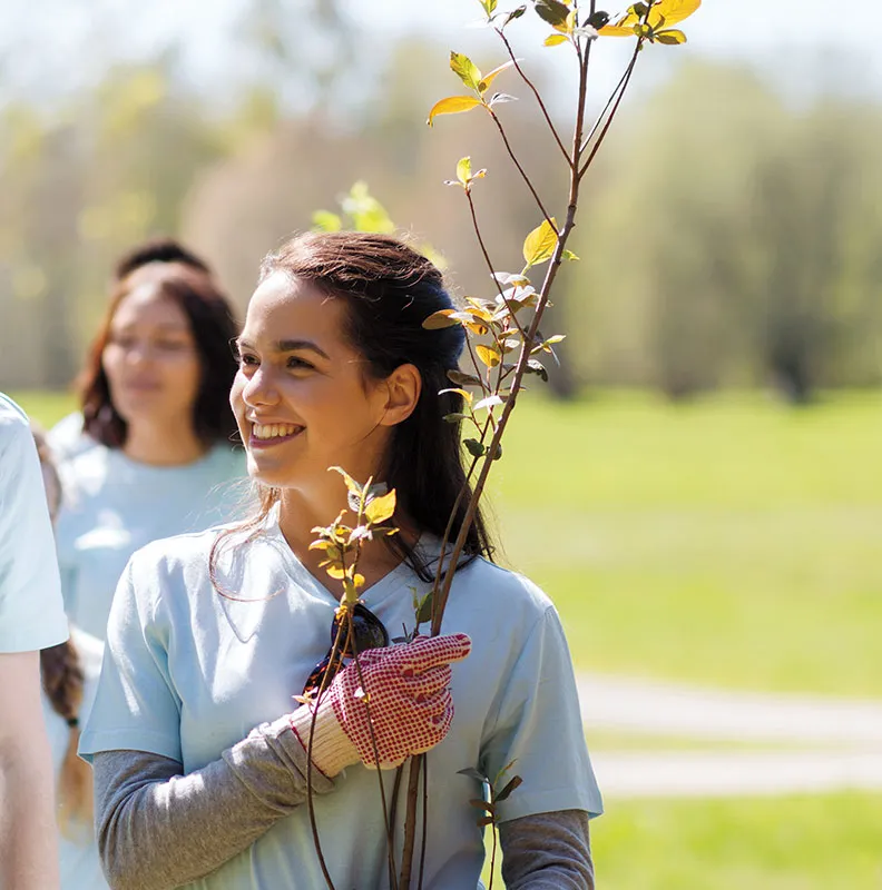 A group of volunteers in light blue t-shirts participate in a tree planting event, one holding young saplings outdoors.