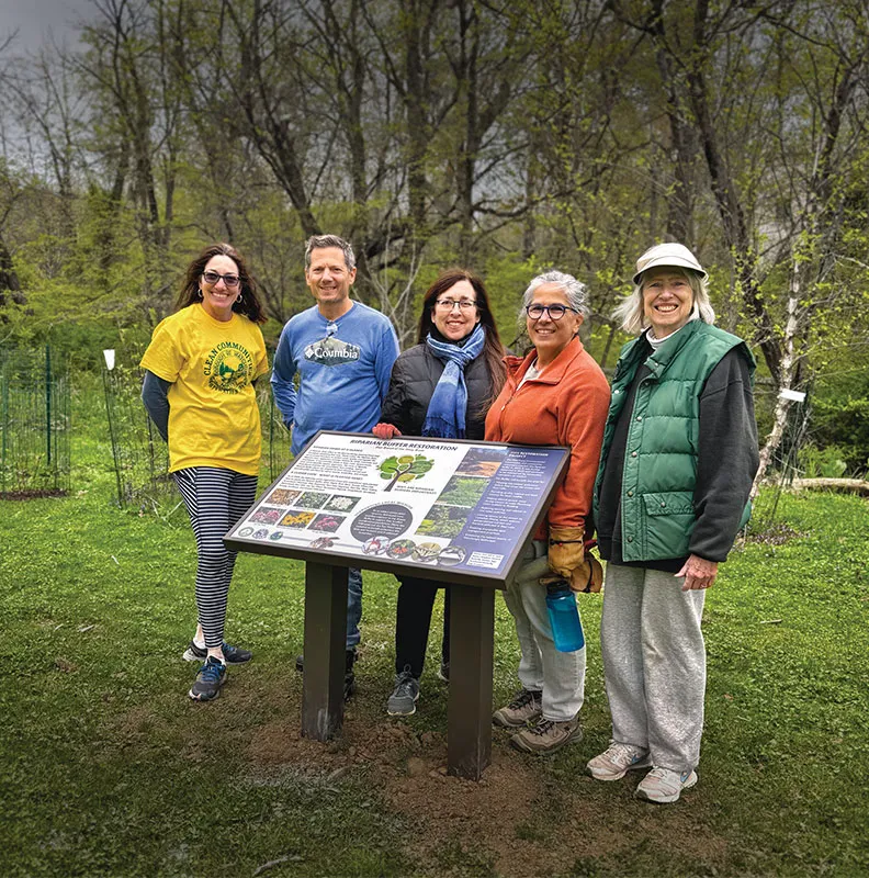A group of five people stands in front of an informational sign about ecological restoration in a lush green area.