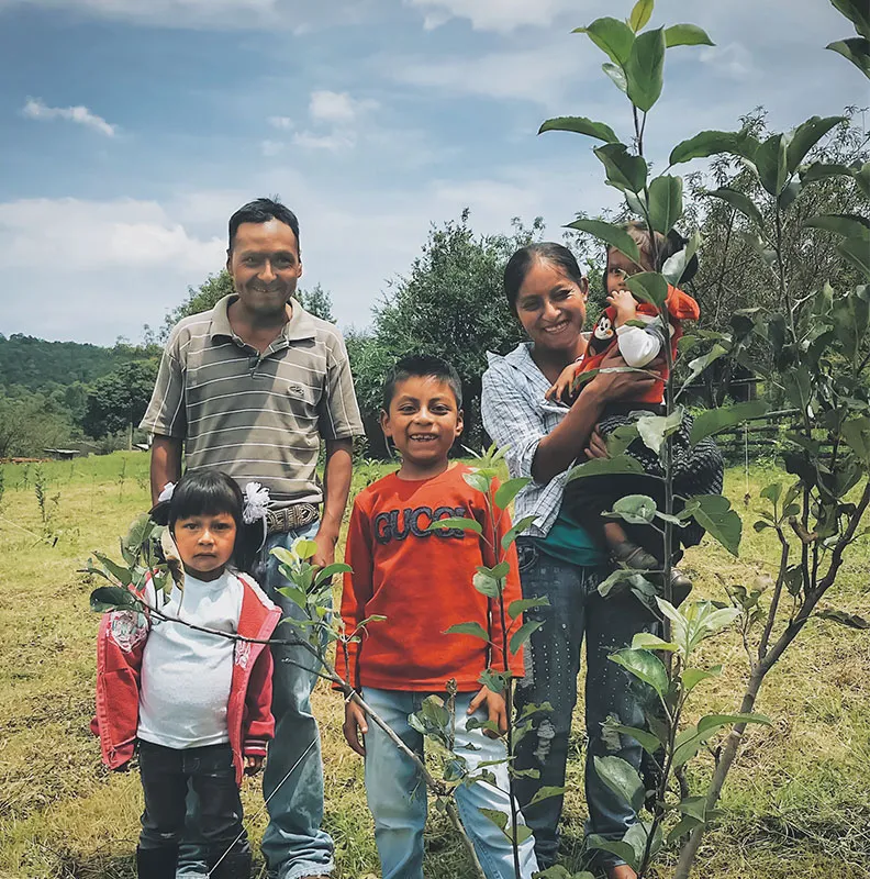 A group of children and adults stand together in a lush green field surrounded by young plants under a cloudy sky.
