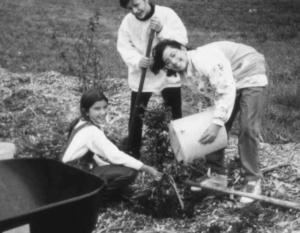 Three children engaged in gardening, with one using a shovel, another pouring water from a bucket, and the third planting seeds.
