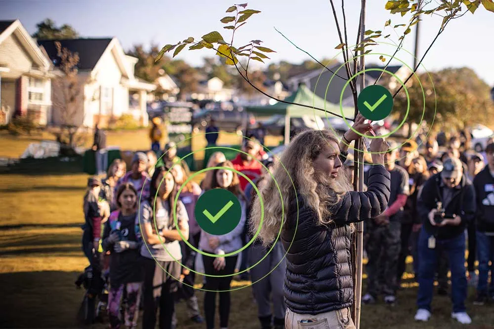 A group of people watches as an individual demonstrates planting a young tree in a community event outdoors.