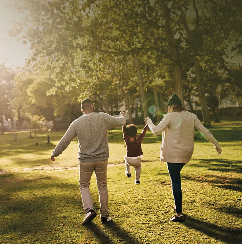 A family walks happily in a sunlit park, holding hands with a child who is playfully suspended between them.
