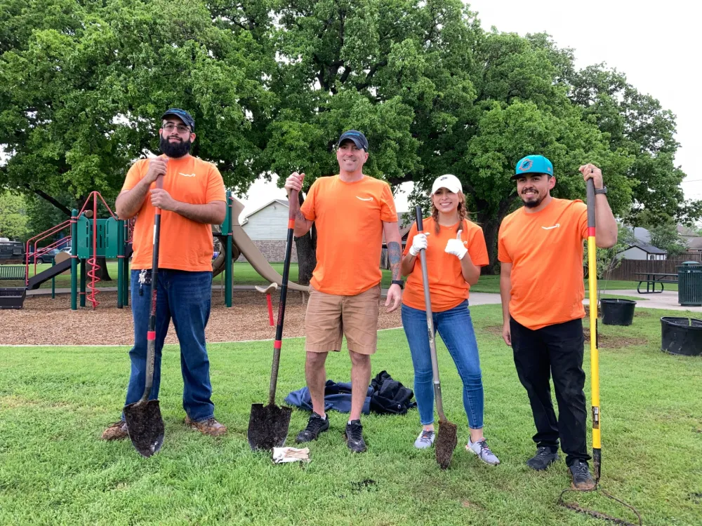 A group of four individuals in orange shirts pose with shovels in a park, surrounded by trees and playground equipment.
