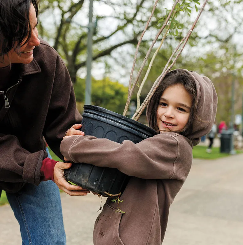 Adult and child outdoors holding a large potted tree together, surrounded by trees and a pathway in a park setting.