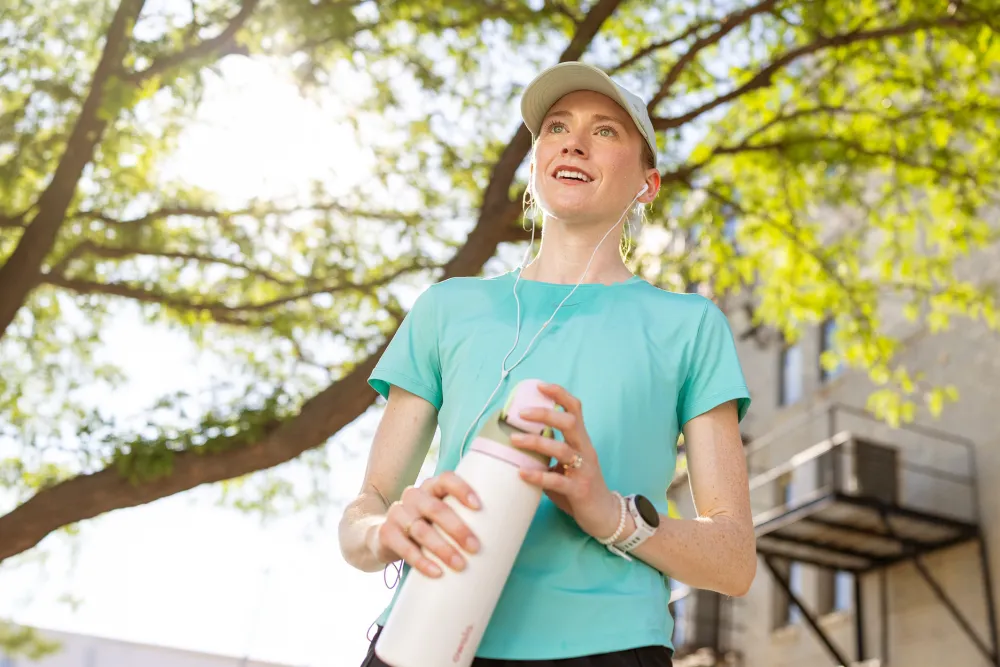 Person in turquoise shirt and cap holding a water bottle outdoors under a tree with sunlit leaves and a building in the background.