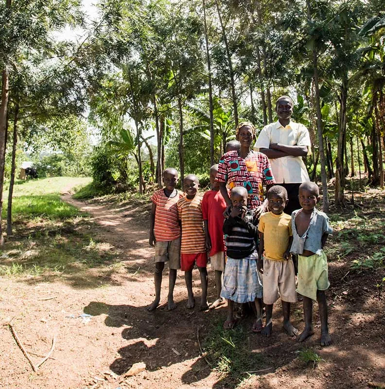 Group of children and two adults standing on a dirt path in a sunny, forested area with green trees surrounding them.