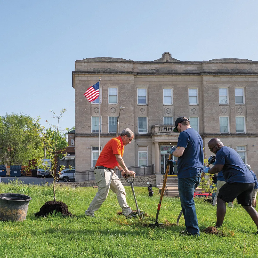 Three men digging holes in a grassy area to plant trees in front of a large building with an American flag.