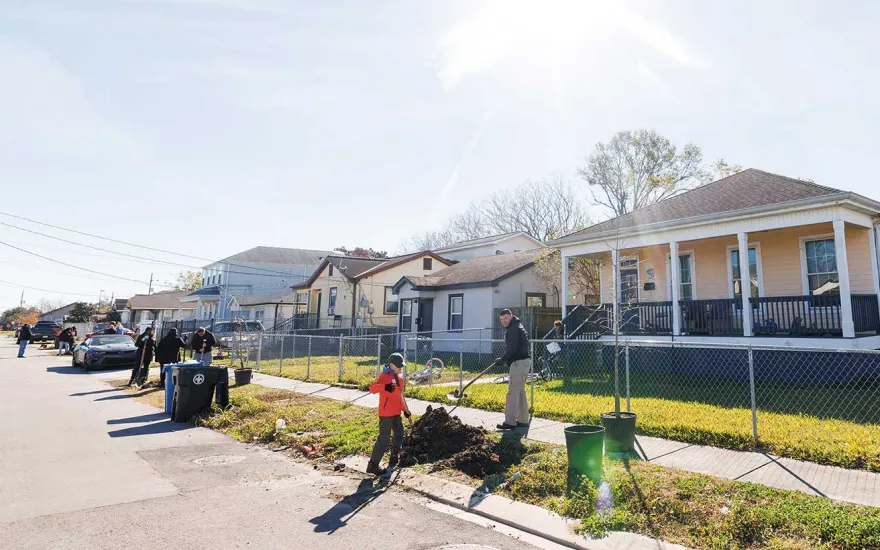 A man and a boy dig a hole in a sunny suburban street, surrounded by neighbors cleaning up yards and parked cars.