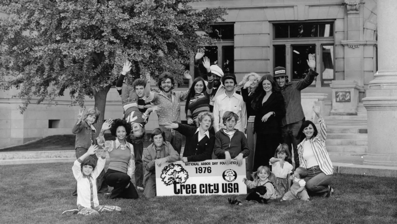 A group of people joyfully poses outdoors with a "Tree City USA" banner, celebrating Arbor Day in 1976, in front of a large building.