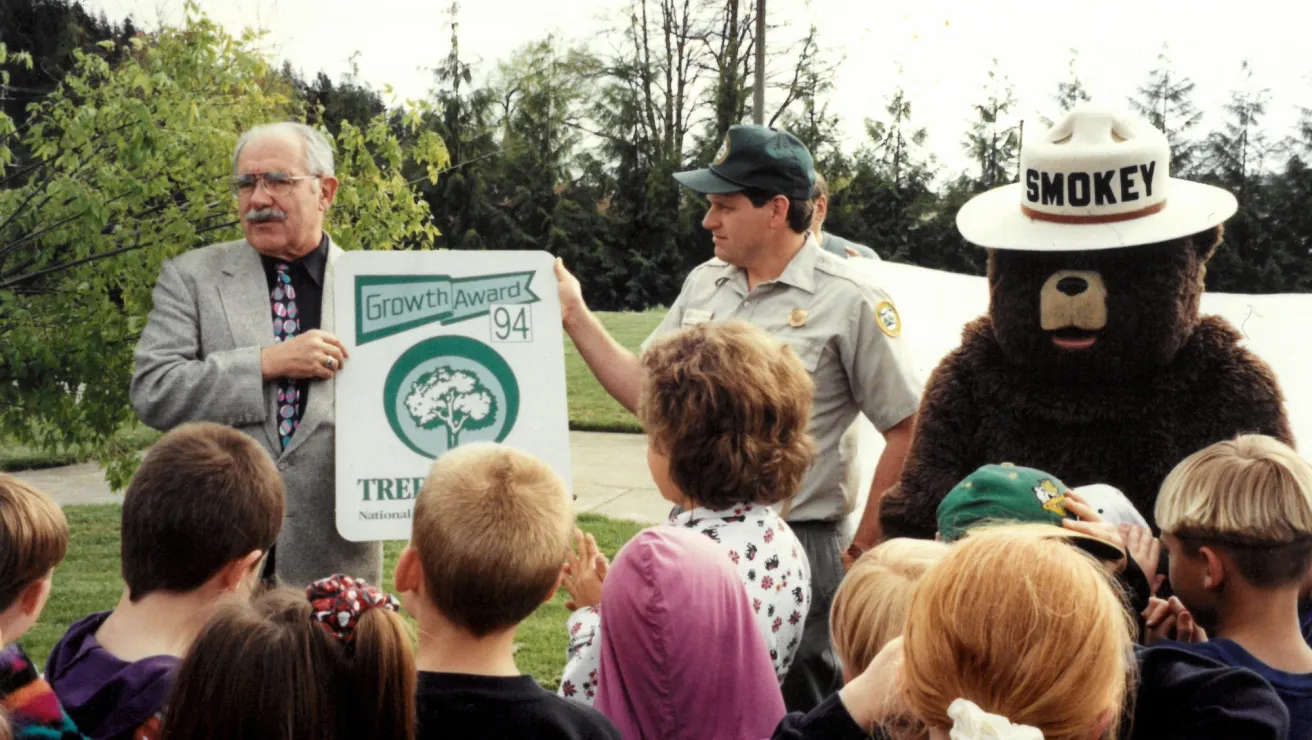 A man in a suit and a park ranger hold a "Growth Award 94" sign beside Smokey Bear. Children gather around, smiling outdoors in a park setting.