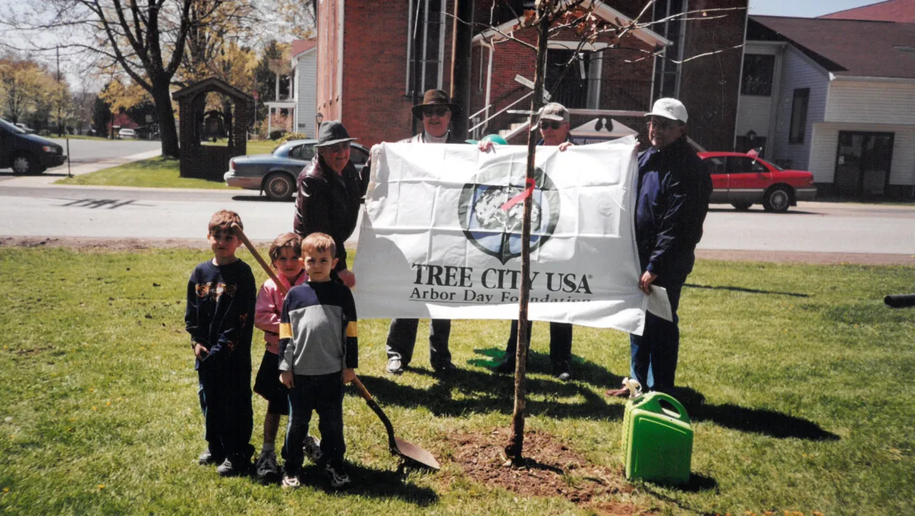 A group of people, including children, stand on grass around a newly planted tree. They hold a "Tree City USA" banner. A sunny day conveys a cheerful tone.