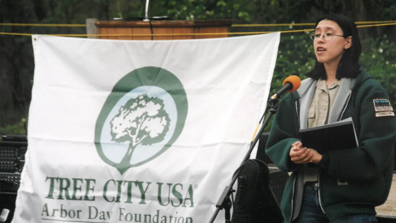 A girl in a green jacket speaks at an outdoor event. They hold a folder and stand beside a "Tree City USA" Arbor Day banner, conveying a sense of environmental awareness.