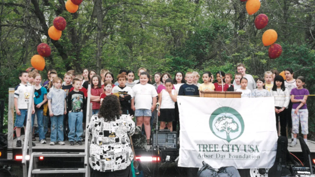 A children's choir performs on an outdoor stage surrounded by trees, with colorful balloons overhead. A "Tree City USA" banner is displayed in front.