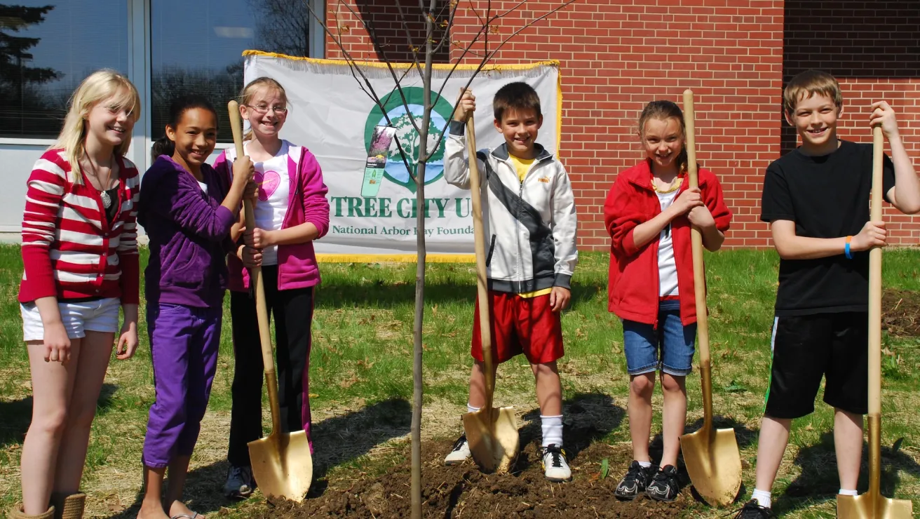 Six children smile while holding shovels around a newly planted tree. A banner reading "Tree City U.S.A." hangs behind them. Sunny, cheerful atmosphere.