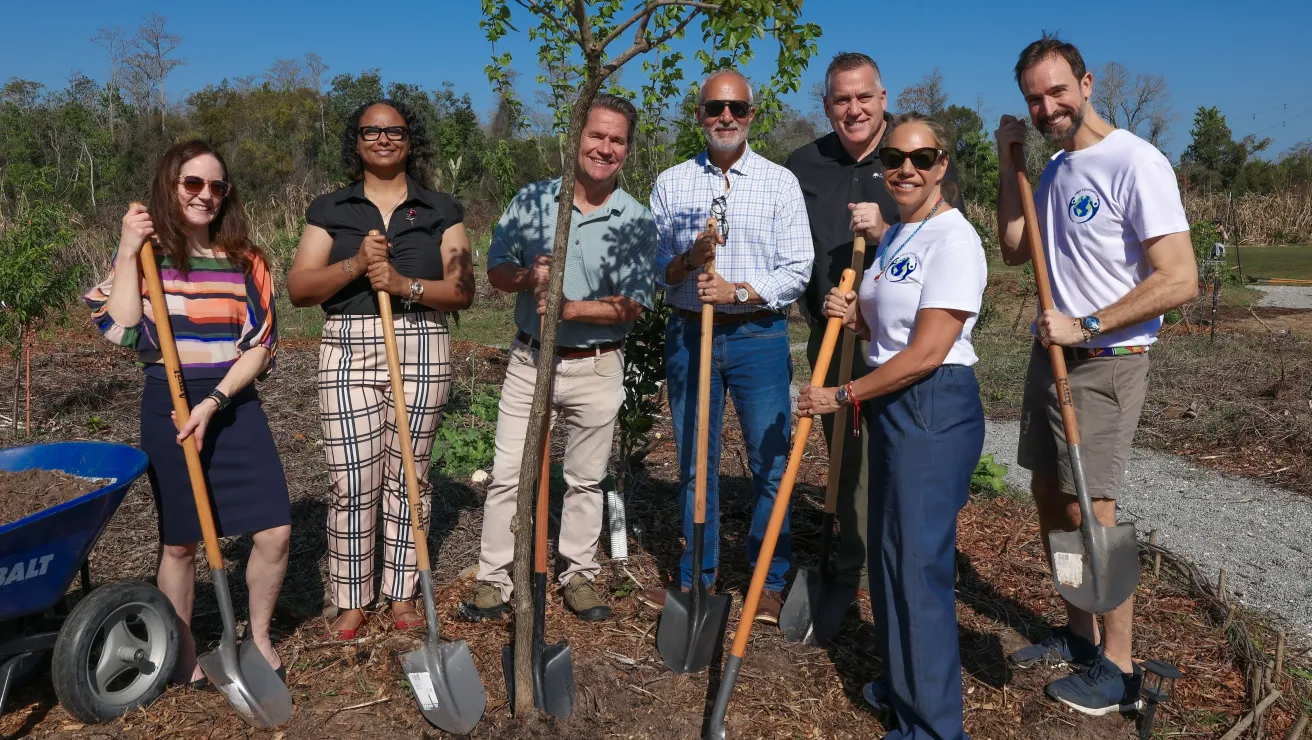 Seven people, including the CEO of the Arbor Day Foundation, stand around a newly planted tree with shovels. 