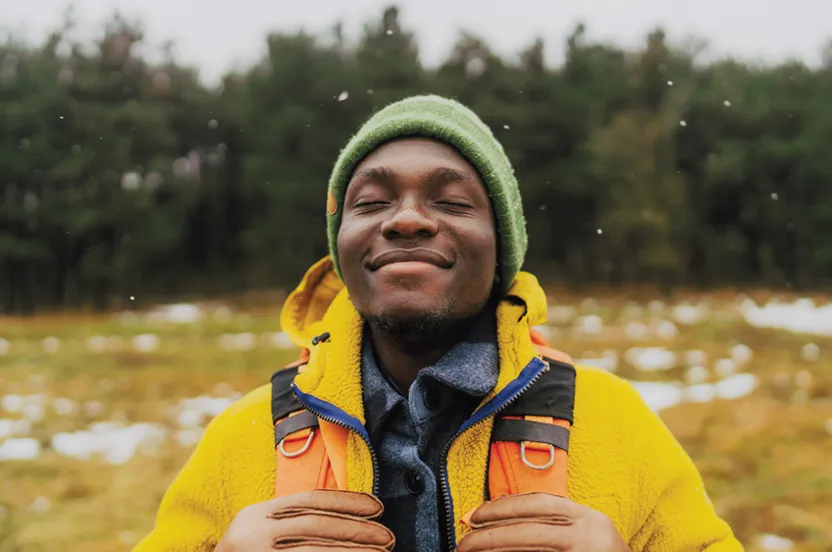 Man smiling with eyes closed, standing outside in front of rows of evergreen trees.