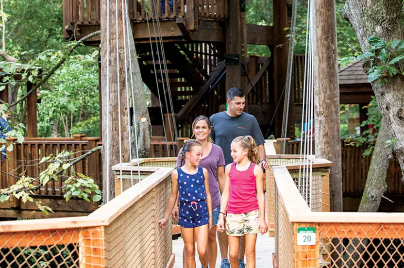 A family walks along a wooden bridge in a forest setting, surrounded by trees and rustic structures in the background.